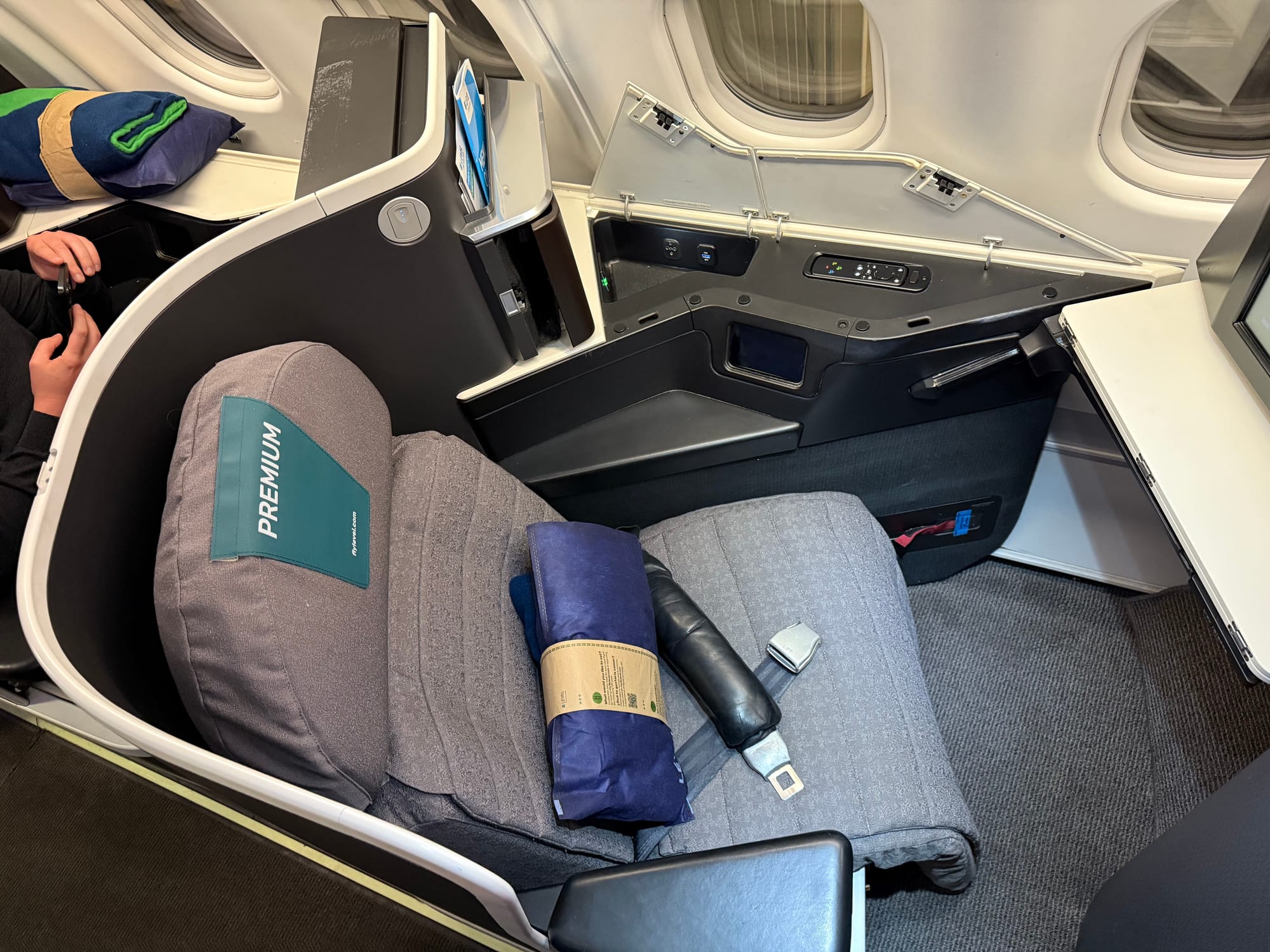 Overhead view of an outward-facing herringbone seat: black and white plastics, grey seat cover, teal headrest cover with "Premium" on it, blue rolled blanket with a paper wrapper.