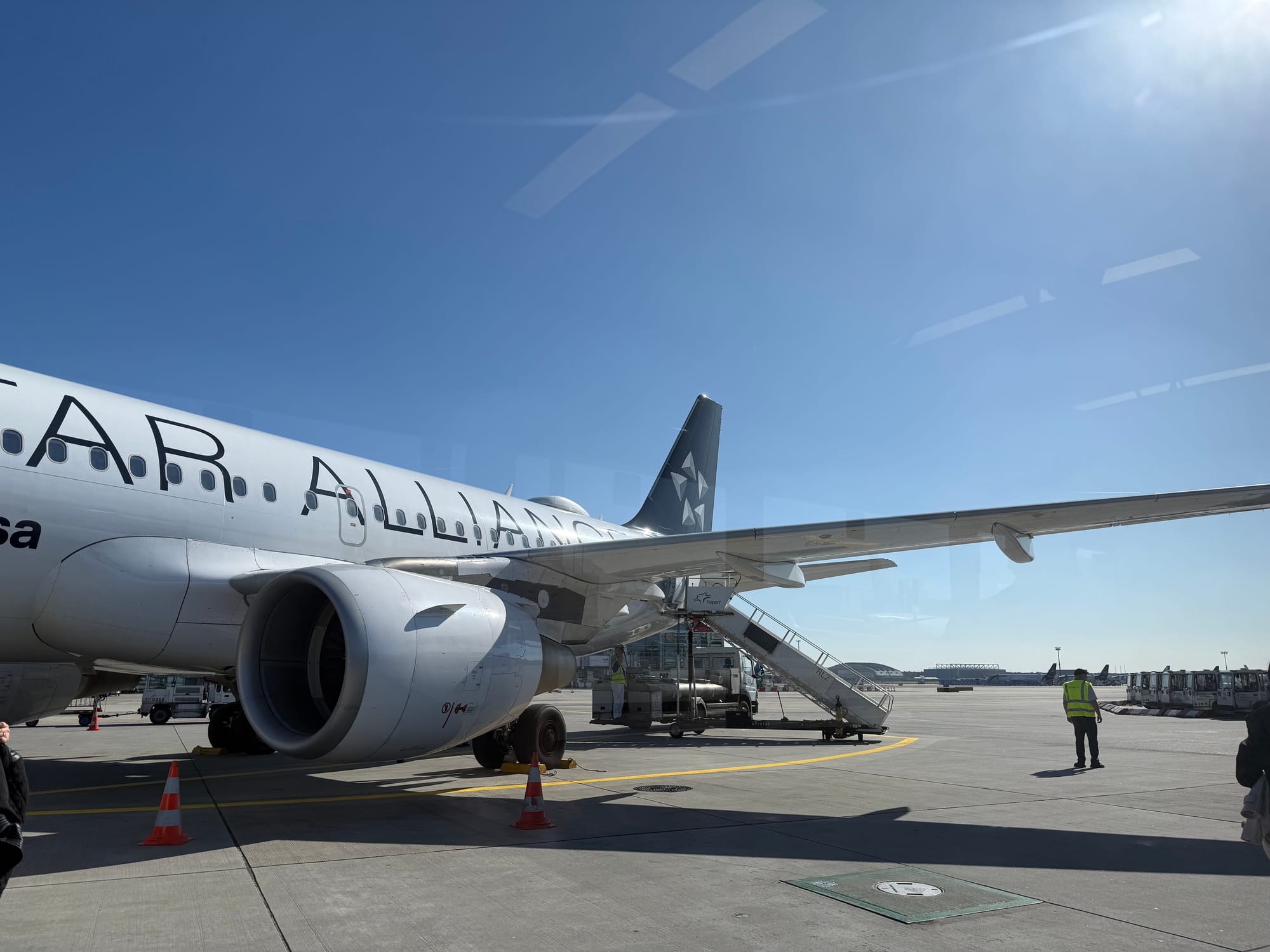 Exterior, airport. An A319 in Star Alliance livery (with the A of Lufthansa's logo visible) sits at a bus gate. Airstairs are connected to the rear door.