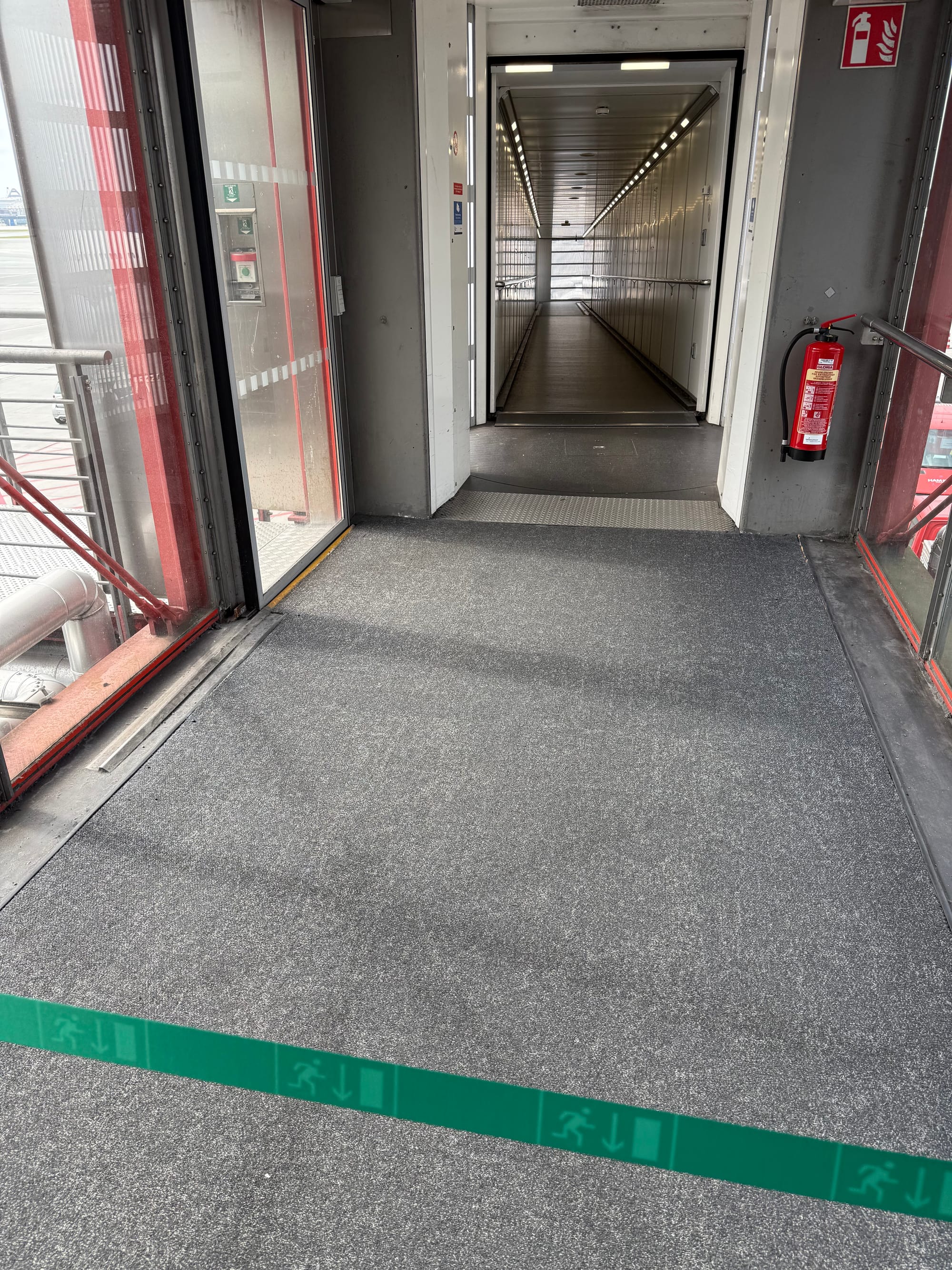 Interior, airport jetway. A tensabarrier-style strip prevents passengers from continuing down towards the aircraft.