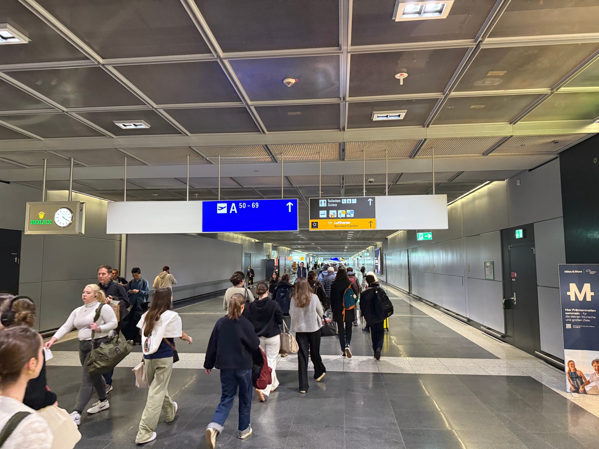 Interior, airport terminal. A long grey corridor extends to infinity with many passengers walking.