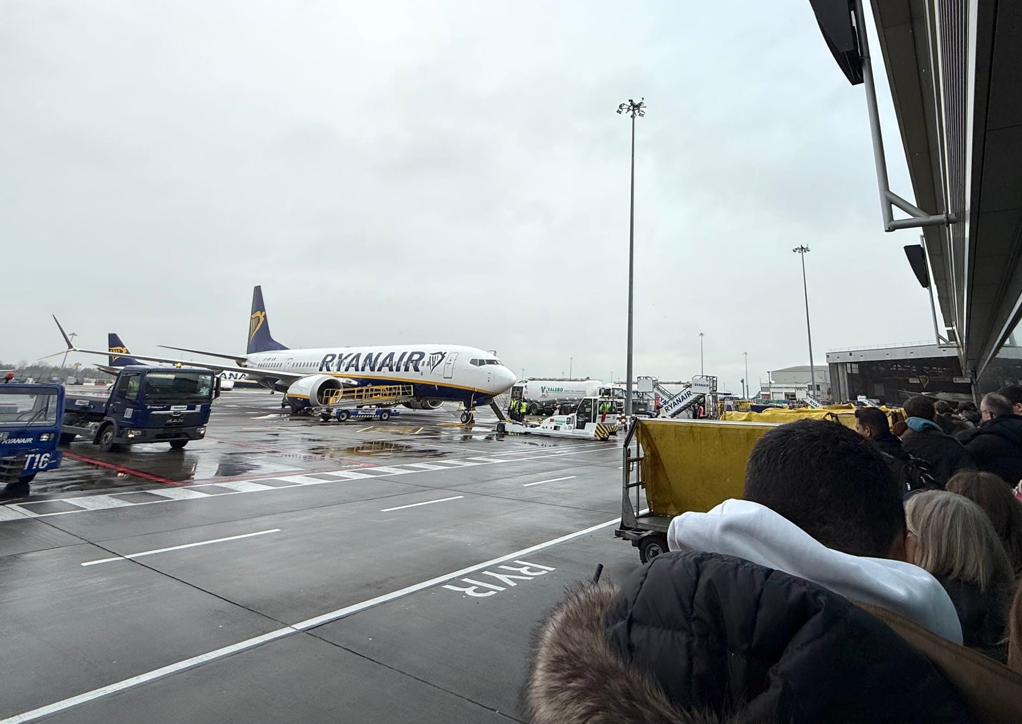 Exterior, airport terminal. Dozens of passengers wait outside the terminal in rainy weather. On the ramp, a Ryanair 737 MAX 8-200 sits without a jetway, with rickety stairs extended to the ground.