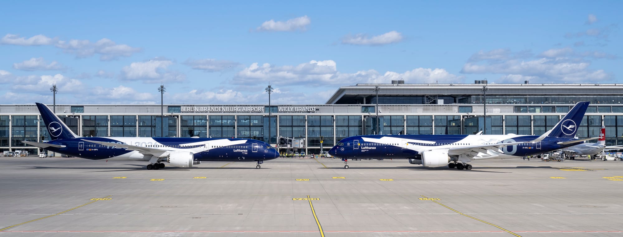 A Lufthansa 787 and A350 in white-crane-on-deep-blue commemorative centenary livery, nose-to-nose in front of Berlin Brandenburg Airport’s terminal