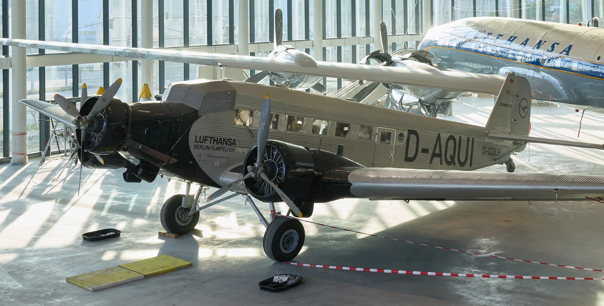 Lufthansa’s Ju 52 in final preparations inside the hangar, showing its Lufthansa crane and “Lufthansa” titles”.
