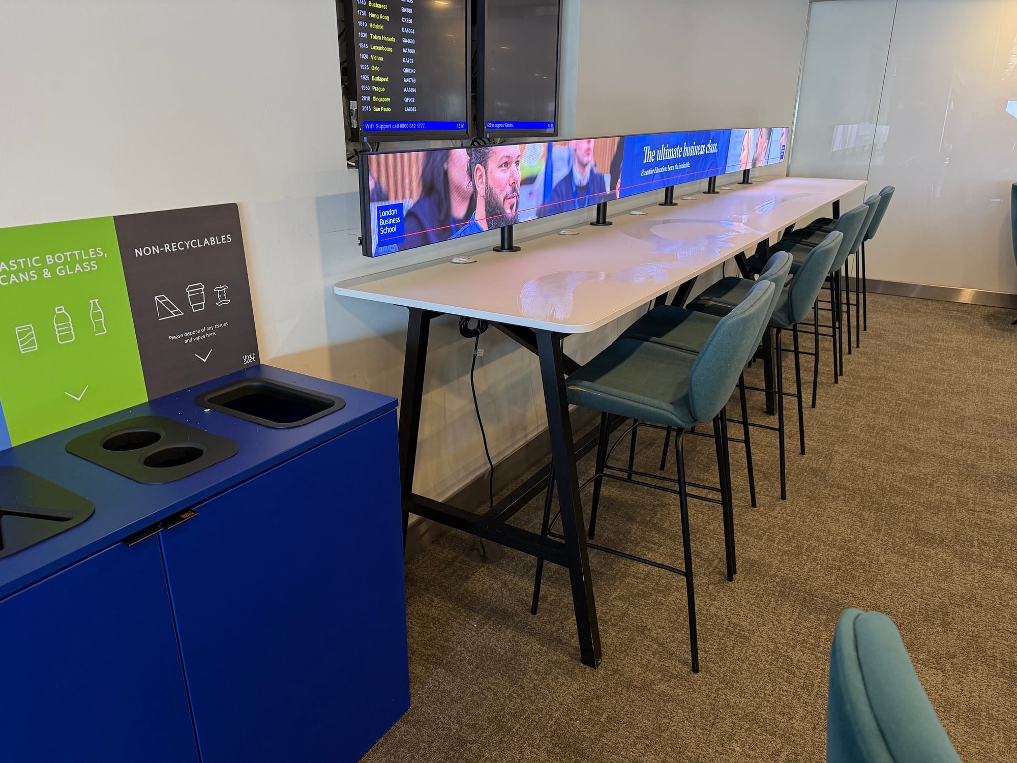 Interior, visibly different lounge. On the left are bins for rubbish and recycling. In the centre and right are six high stools with low backrests at a single high table. On the table’s full length there is a very wide screen showing an ad for a business school.