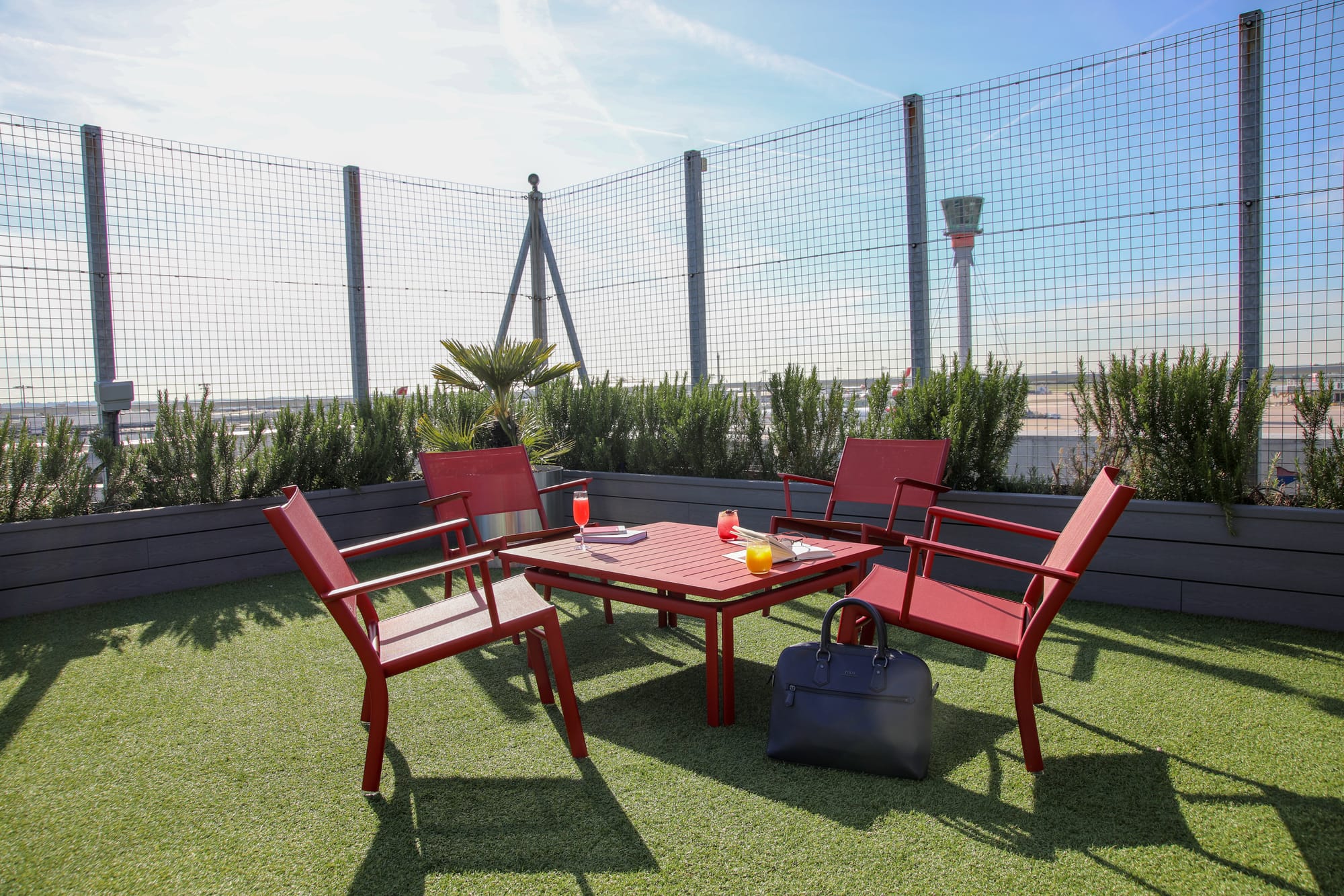 Rooftop photo: an artificial turf area is contained by grey planters with rosemary plants, with cage-type fencing behind. In the distance is the iconic Heathrow control tower.