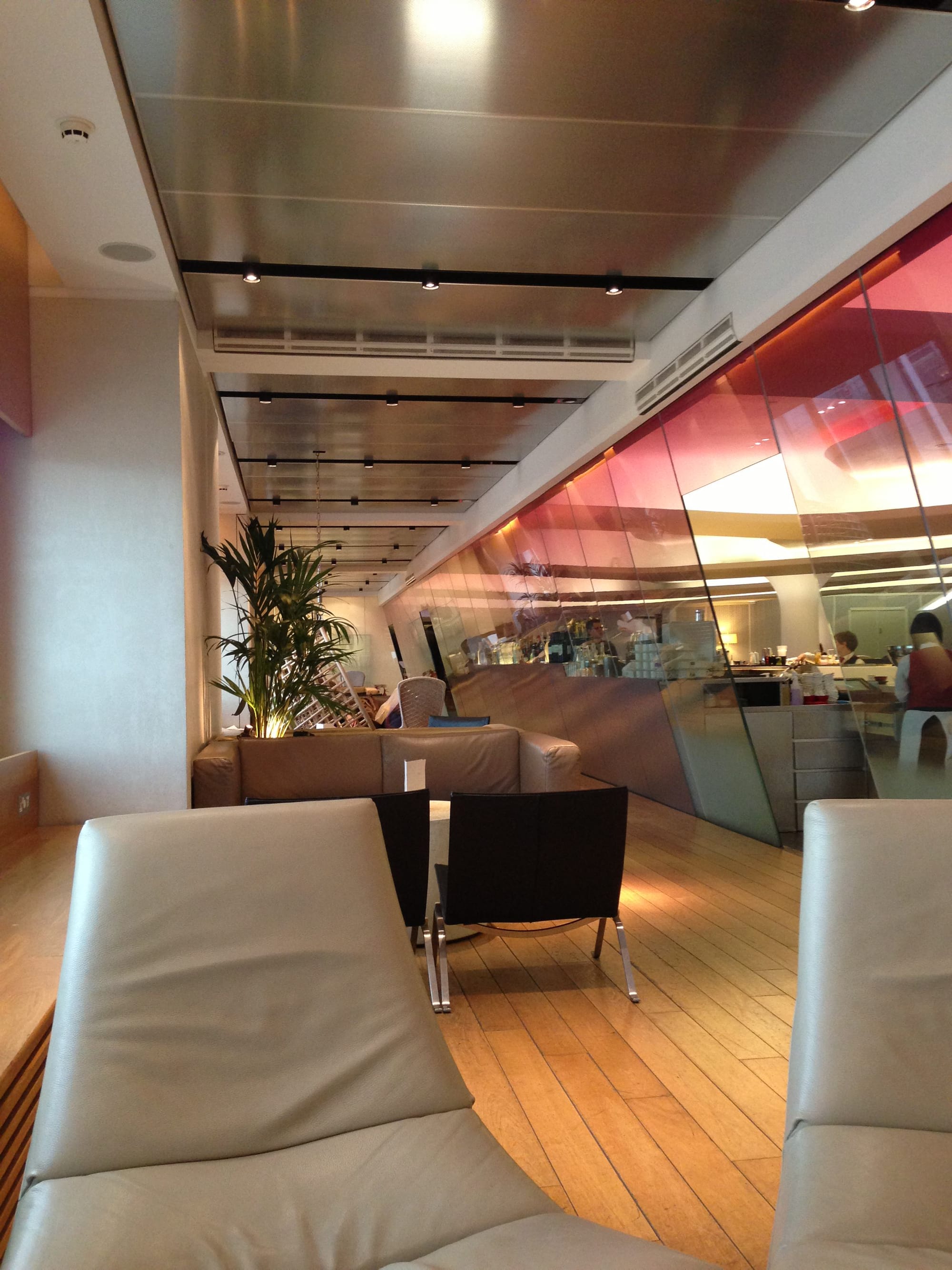 Interior, airline lounge — same lounge, same period, relatively low-res photo from the early 2010s. Low chairs in grey and black sit inside a similar space to the previous photo, with the pink-tinged glass panels now angled towards us.