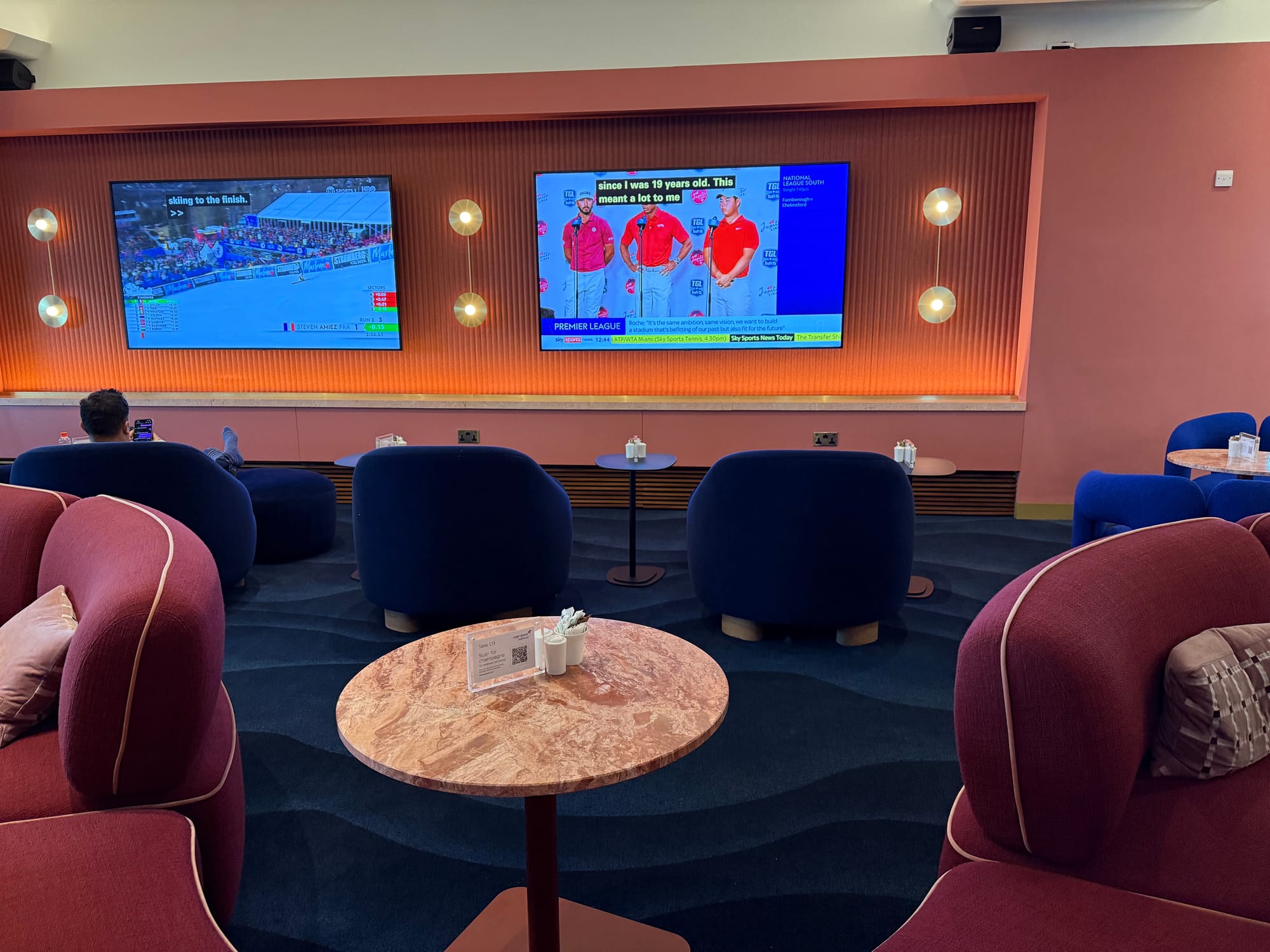 Interior, lounge. Two TVs, approx 75-85”, show sports. In front of them are some blue chairs and behind those are red S-shaped sofas.
