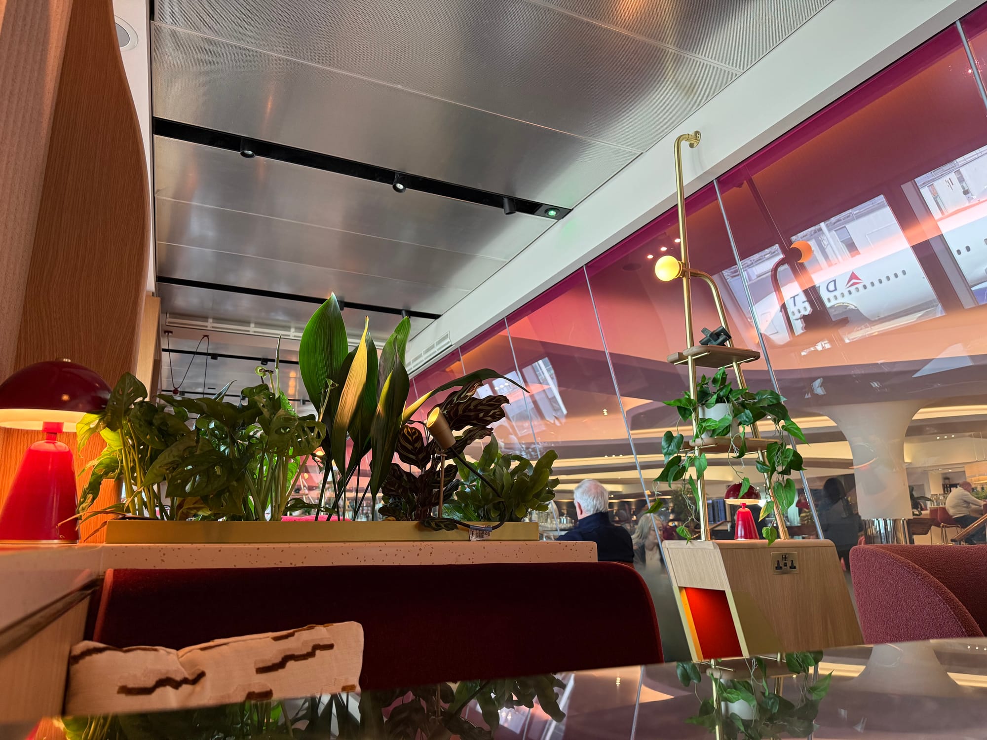 Closeup, same lounge: the rose tinted glass on the right, then the lighting/power/side table elements, with planters as soft dividers, viewed from a booth.