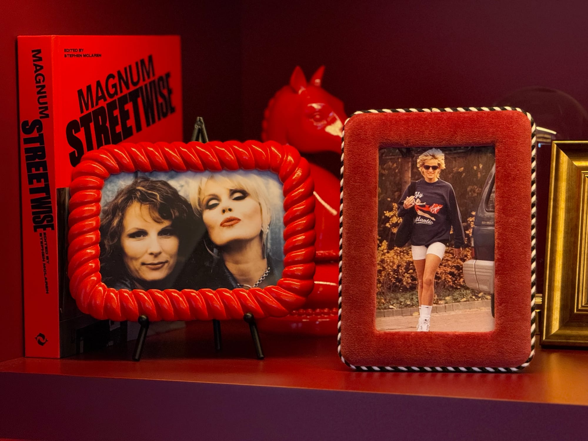 Closeup on a bookshelf wall. Red book, “Magnum Streetwise”. Photo in red frame, Jennifer Saunders and Joanna Lumley as their Absolutely Fabulous characters. Red horse bookend. In different red frame, the well-known photo of Princess Diana wearing the blue longsleeve Virgin Atlantic sweatshirt.