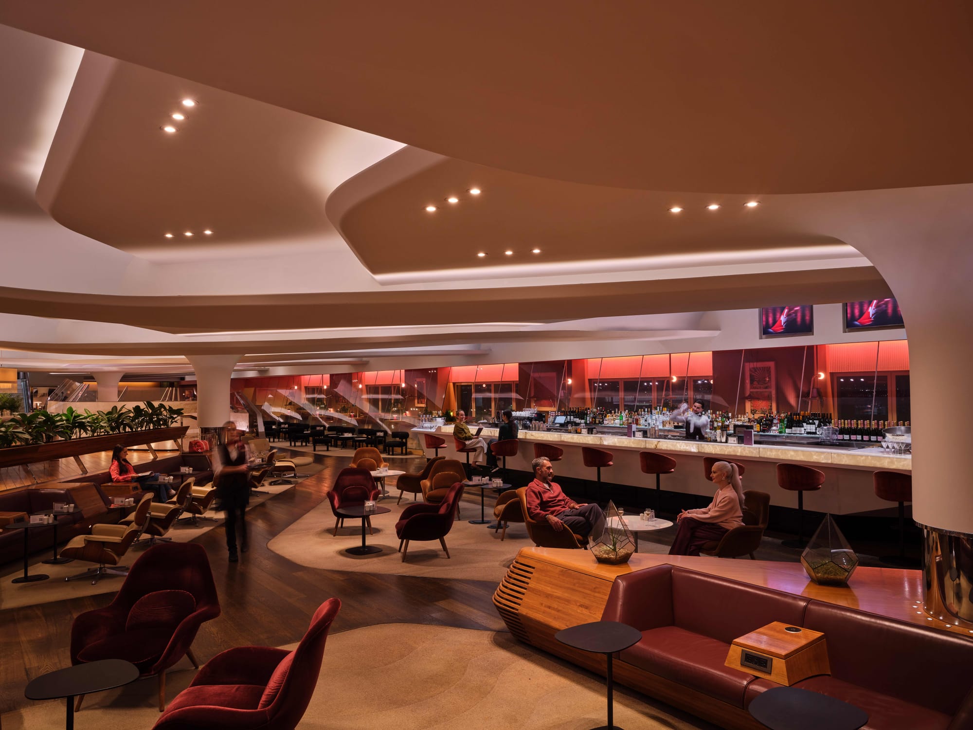 Interior, business class lounge, nighttime. Red aesthetic and moodlighting at the back, swoopy 1960s modernist ceiling, a few people scattered around on mostly red chairs, a big bar with bar staff in the middle.