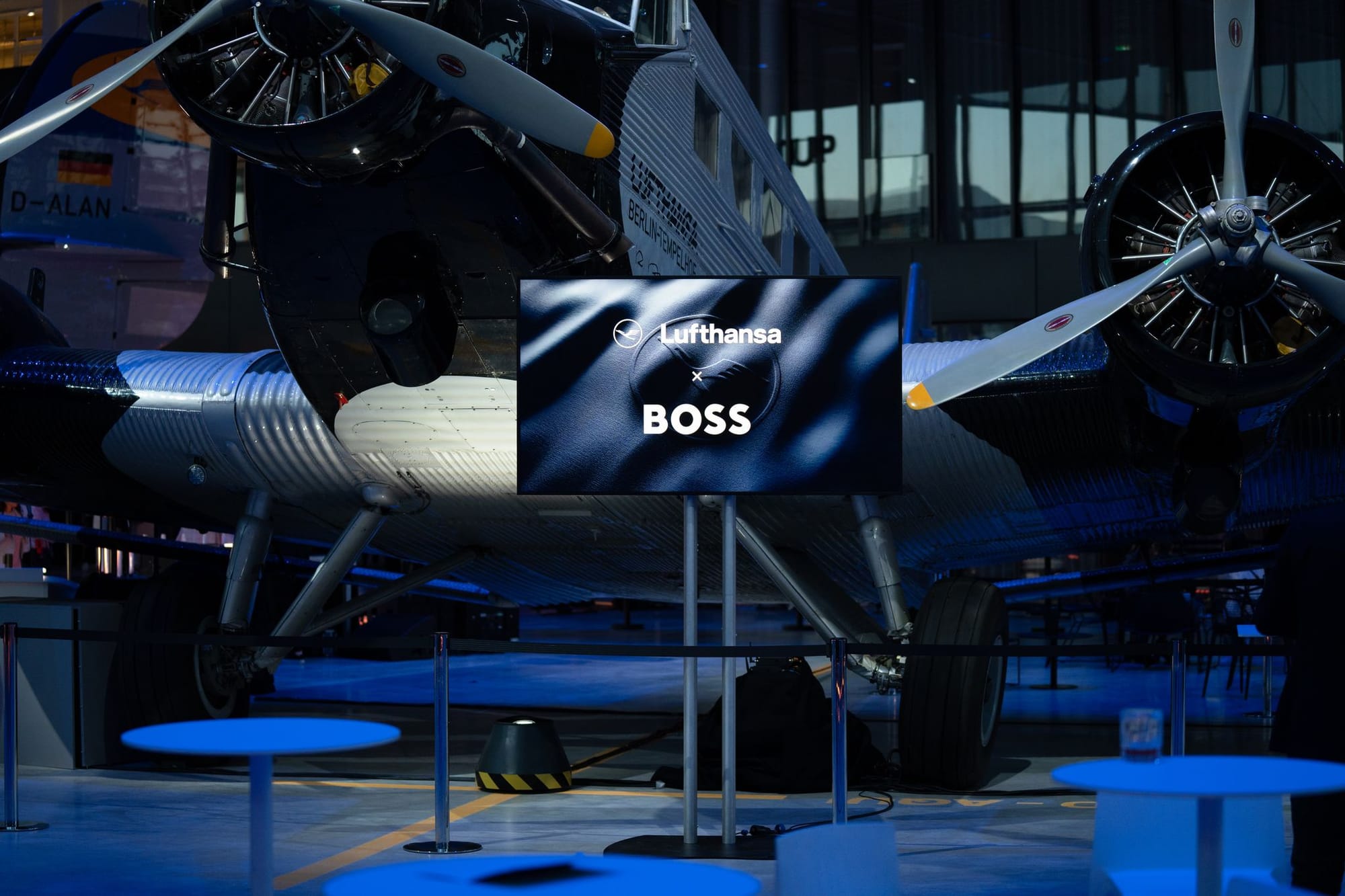 Event with tables ready for attendees, with a large flat-panel screen showing [Lufthansa logo] x [BOSS logo] in front of a Ju 52 with modern Lufthansa markings.