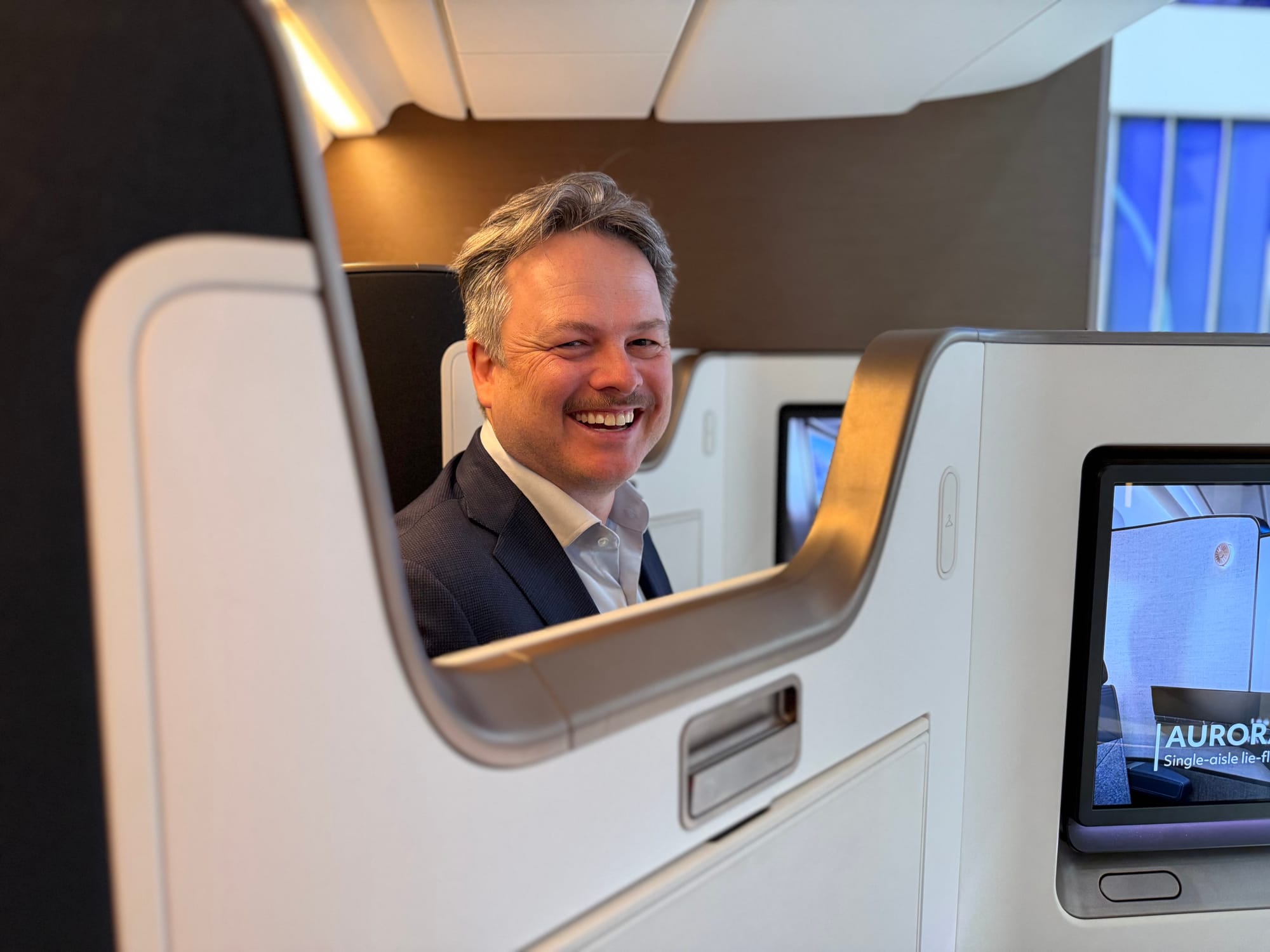 bc A man smiles back through a “chatting gap” element on the Collins Aurora aisle-facing herringbone seat inside a trade show stand. 