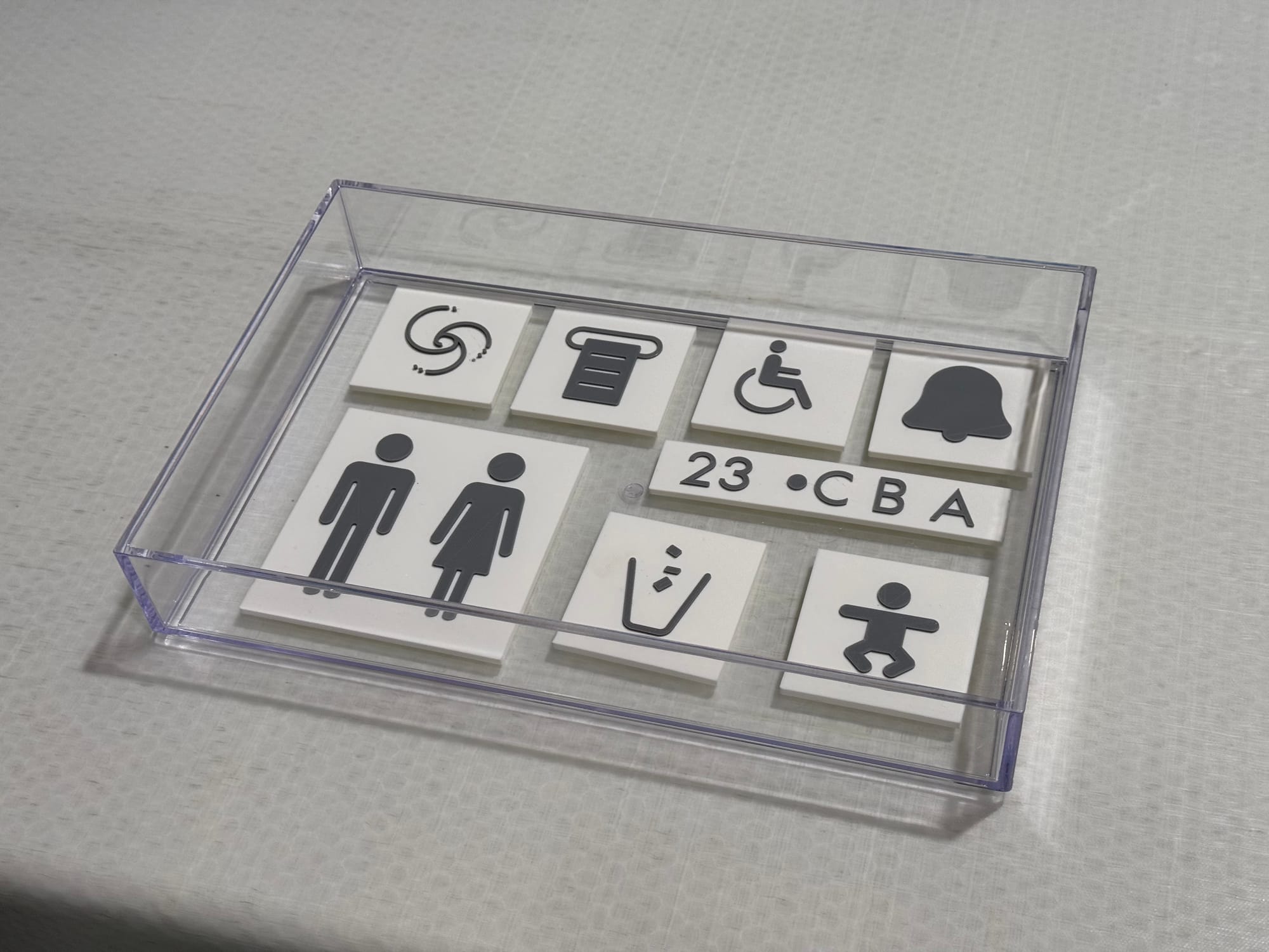 A series of raised placard signage elements, including those for standard male-with-legs and female-in-dress iconography, a trash bin, small baby —&nbsp;plus letters and numbers, and others for a bell, with one being a sort of swirling series of lines with dots at the end of them.