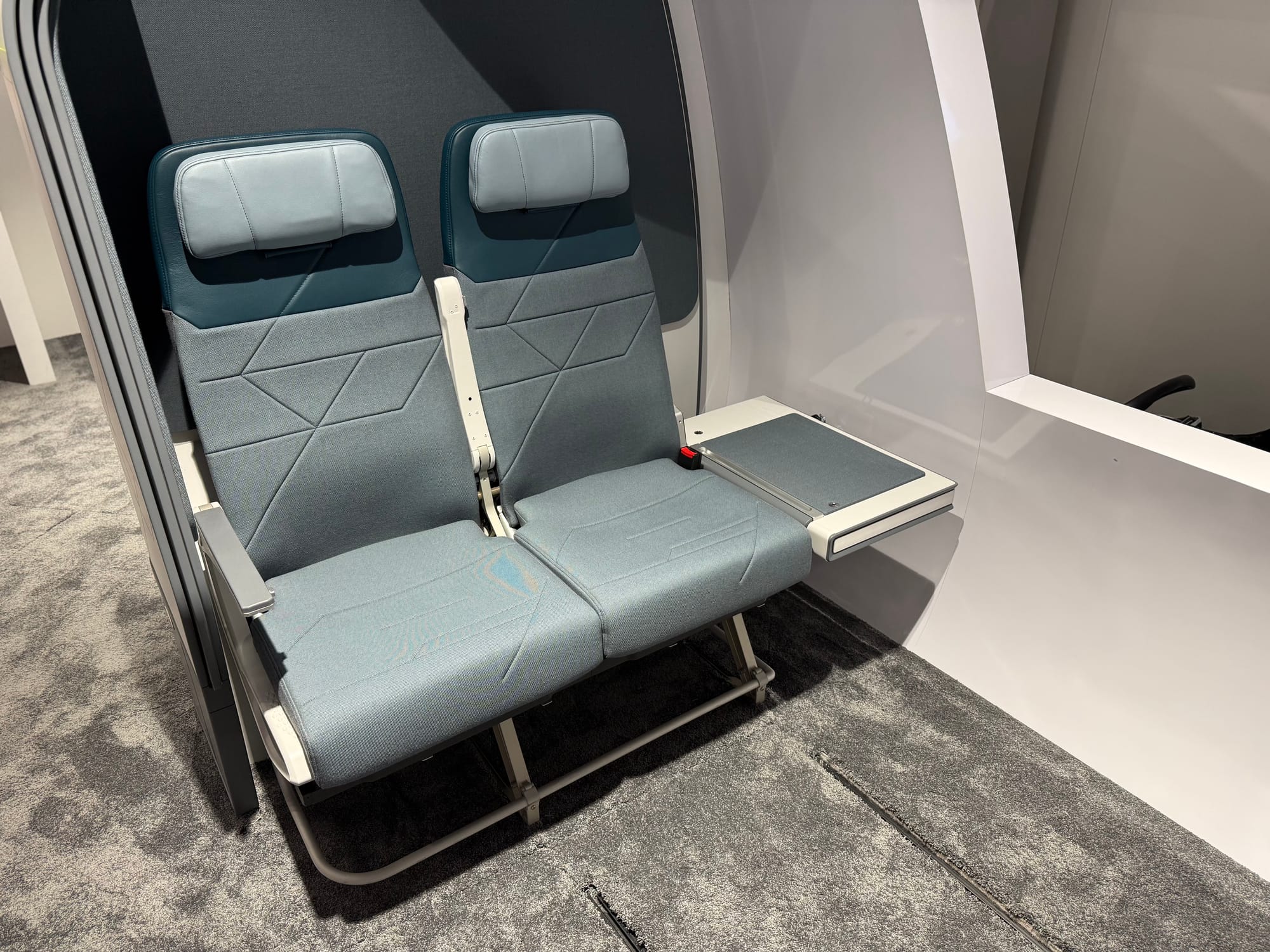 Collins SkyNook mockup. Two seats are at the rear of a fuselage mockup area. Next to what would be the middle seat, replacing a window seat, is a folded up table about 2/3 the width of the seat pan. Underneath is space that would fit a labrador-sized dog.