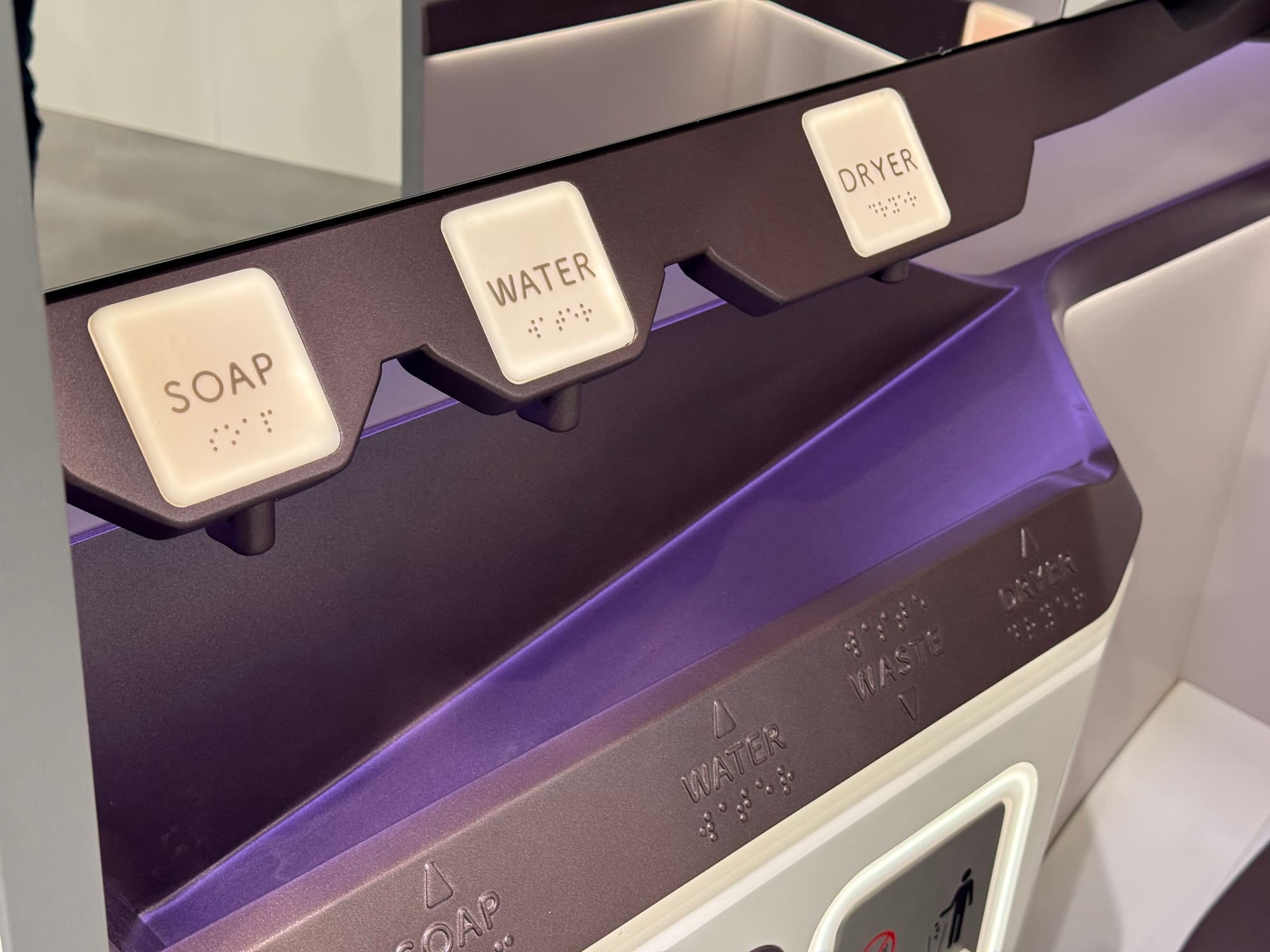 Closeup on the soap, water, waste and dryer functions in a lavatory. There are multiple signs on them, using raised lettering and braille. Above the sink, the soap, water and dryer ones are backlit in white squares.