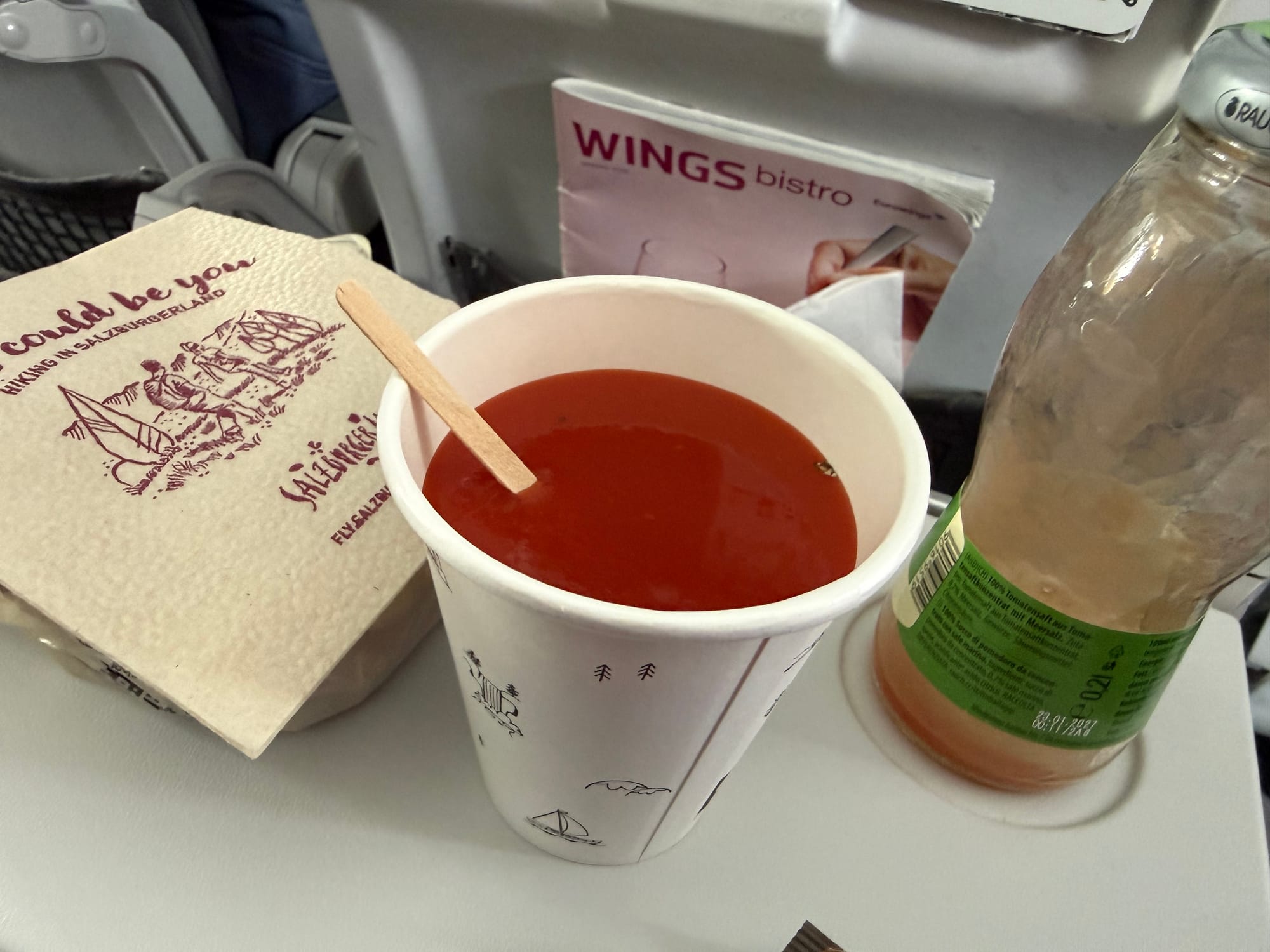 A sandwich in a bag hidden by a napkin, and a paper cup of tomato juice.