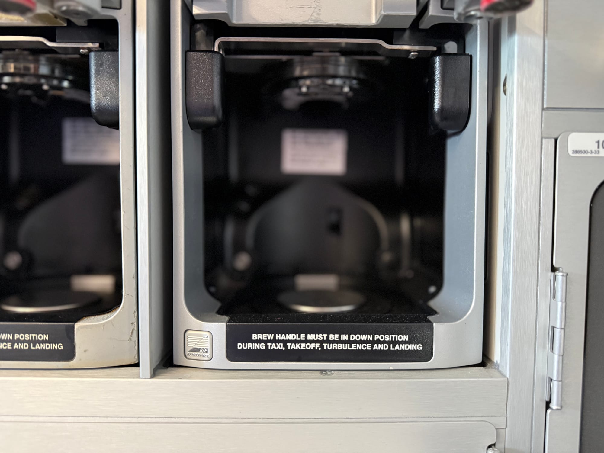 Closeup on coffeemakers. One has a B/E logo. The other is quite heavily dinged.