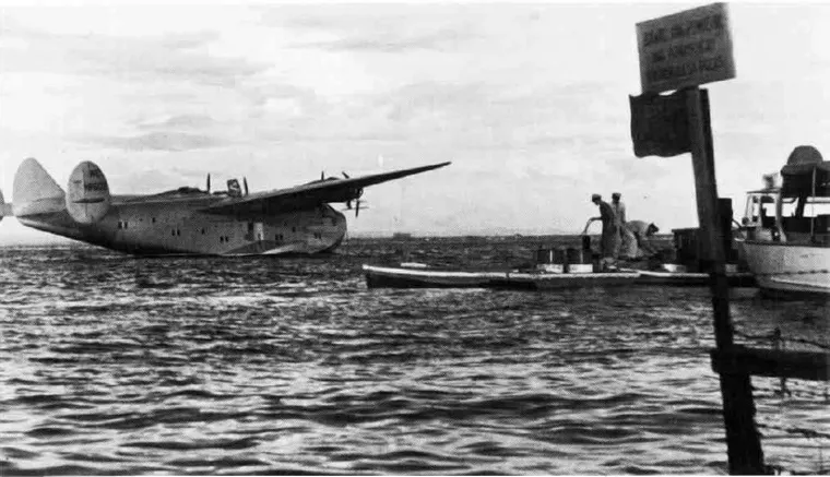 a large flying boat sits on the water. in the foreground two men are preparing to clean her from a jetty