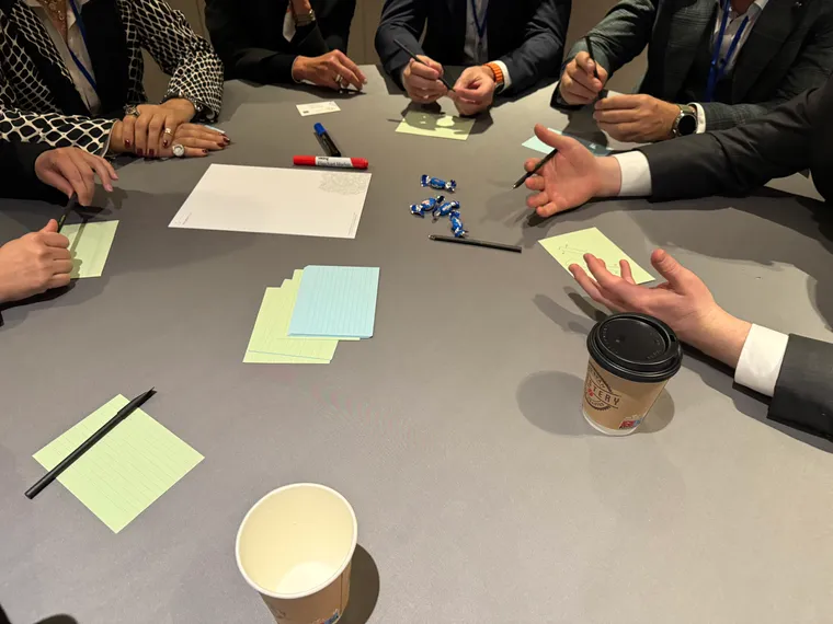 People sit around a table with coffee cups and index cards. We only see their hands. One appears to be speaking with their hands.