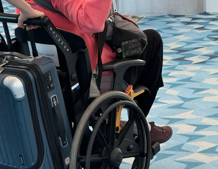 A person in an airport wheelchair, holding a metal walking stick, is wheeled by an assistant.