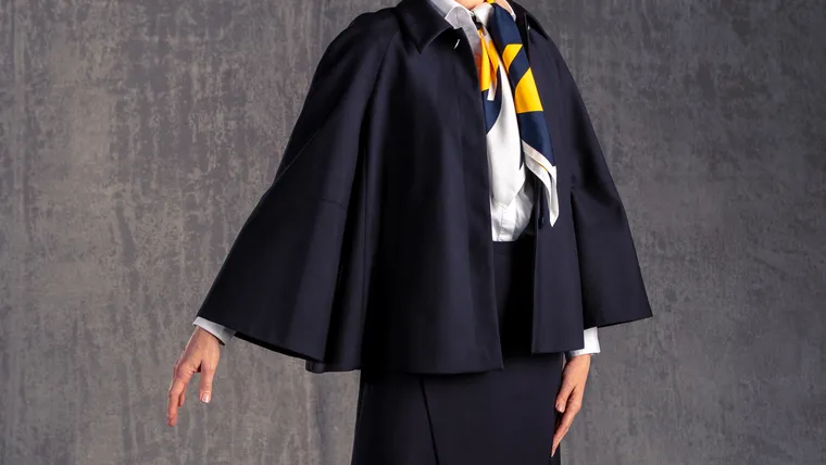 A woman wearing a dark blue cape and blue-white smart business uniform, with a blue-white-gold scarf, lifting one hand