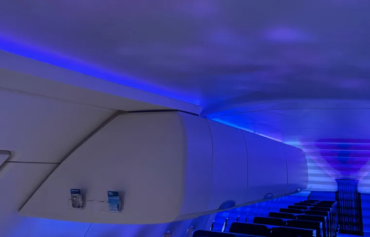 Interior, trade show mockup. Large bins lit with LED mood lighting are inside a small narrowbody mockup cabin, with a screen showing many rows behind.