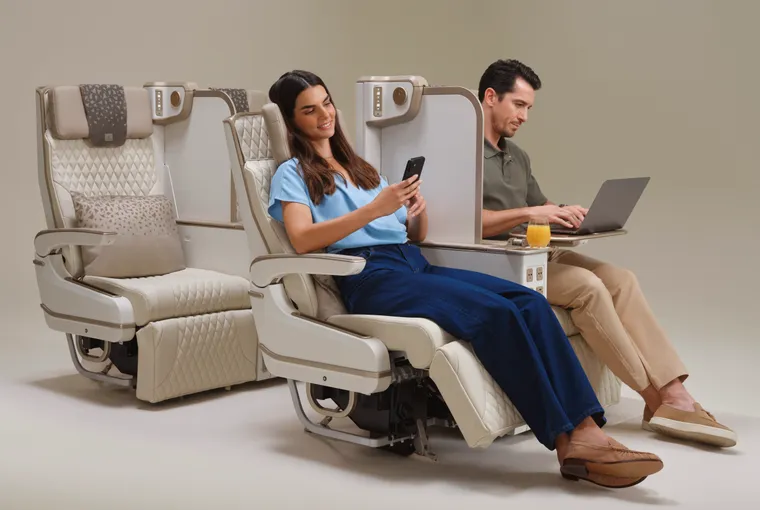 Seat mockup. A man and woman sit in beige premium economy seats. She is reclined, he is working on a laptop. A privacy divider is between them.