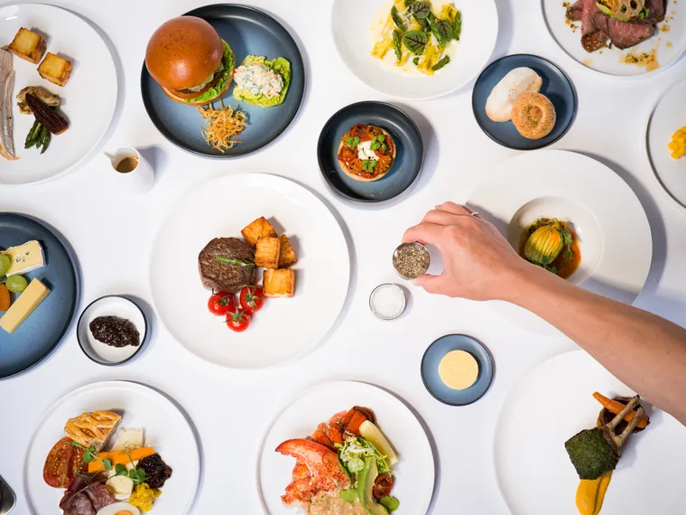 A series of mostly plain white plates and some dark blue gradated plates with food on them, shot from above.