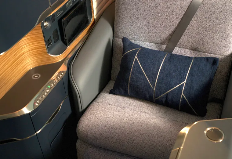 Closeup on a first class seat with grey fabric, grey leather, and blue/wood surrounds. There is a blue cushion with angular design reminiscent of the BA Speedmarque brand element.